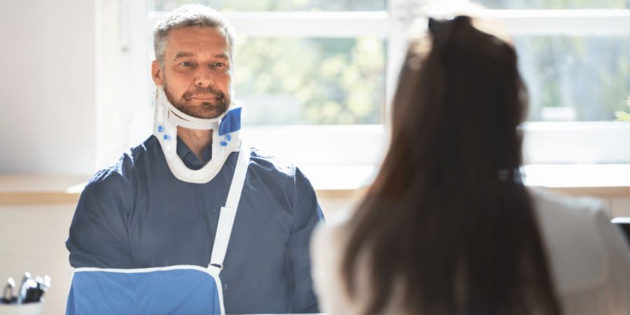 Man in neckbrace with arm in sling at desk with doctor.