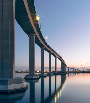 Twilight view of a curved, elevated bridge with streetlights reflecting on calm water below