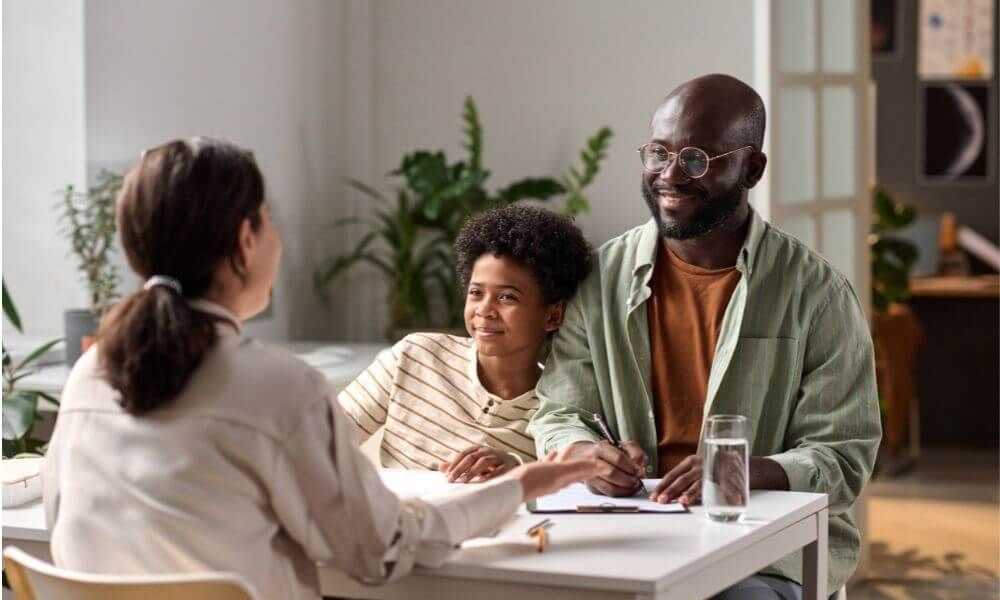 A man and a young child smiling, sitting at a table during a meeting with a woman in an indoor, plant-adorned setting.