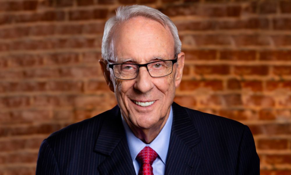 Elderly gentleman in a suit smiling against a brick wall background.