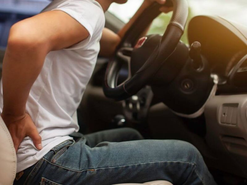 A person in a white shirt sits in a car, gripping the steering wheel while wincing and holding their lower back, suggesting discomfort in the vehicle's interior.