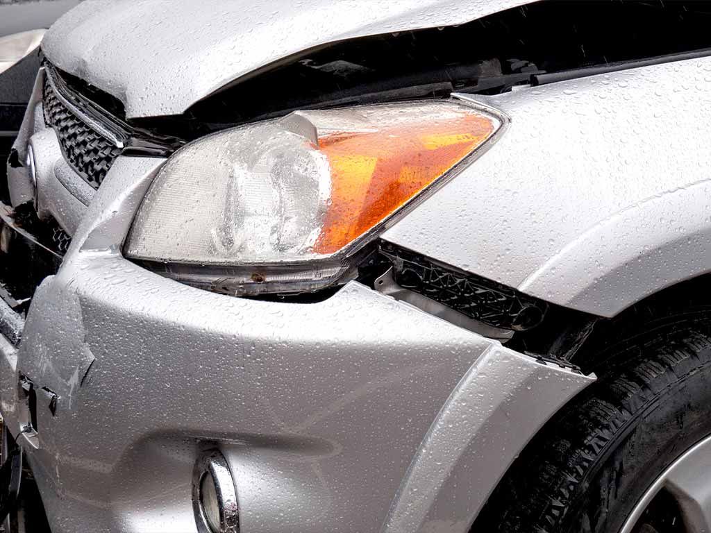 Close-up of a damaged silver car with a dented front bumper and headlight, covered in raindrops.