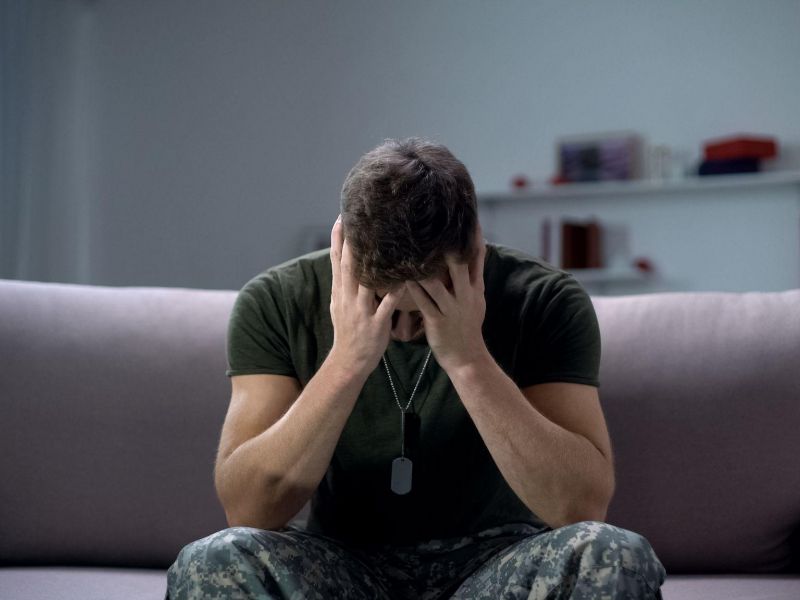 Man in military uniform sitting on sofa, hands covering face, representing stress or anxiety in a domestic setting.