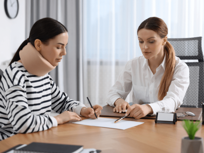 A woman with a neck brace sits at a table, writing while another woman in a white shirt points to paperwork, both engaged in discussion in a bright, modern office setting.