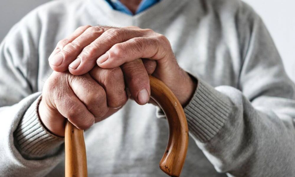 Close-up of an elderly person's hands resting on a wooden cane, symbolizing the support and stability provided by Supplemental Security Income (SSI) services. The person is wearing a grey sweater, indicating a sense of comfort and security in their daily life.