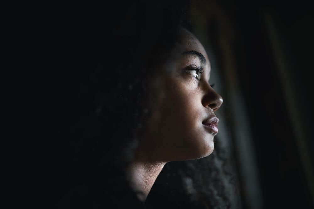 Profile of a thoughtful woman looking out a window, with soft light illuminating her face against a dark background.