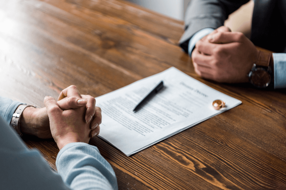 A document with text lies on a wooden table, while two hands clasped in contemplation face each other, suggesting a serious discussion or negotiation in a professional setting. A pen rests nearby.