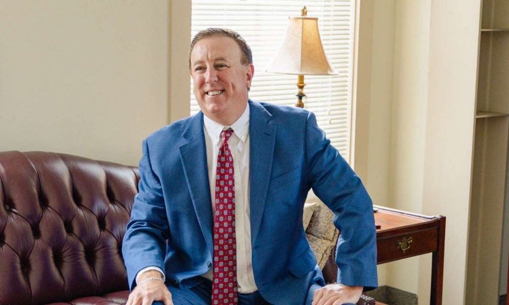 Smiling man in a blue suit sitting on a couch in an office with a lamp in the background.