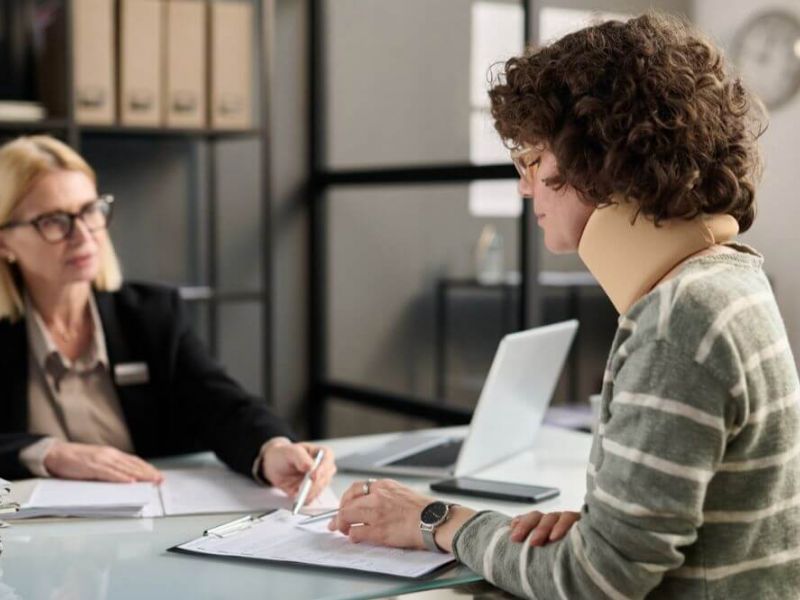 Two people sit at a desk, one signing a document; in an office with shelves and a laptop, suggesting a professional setting.