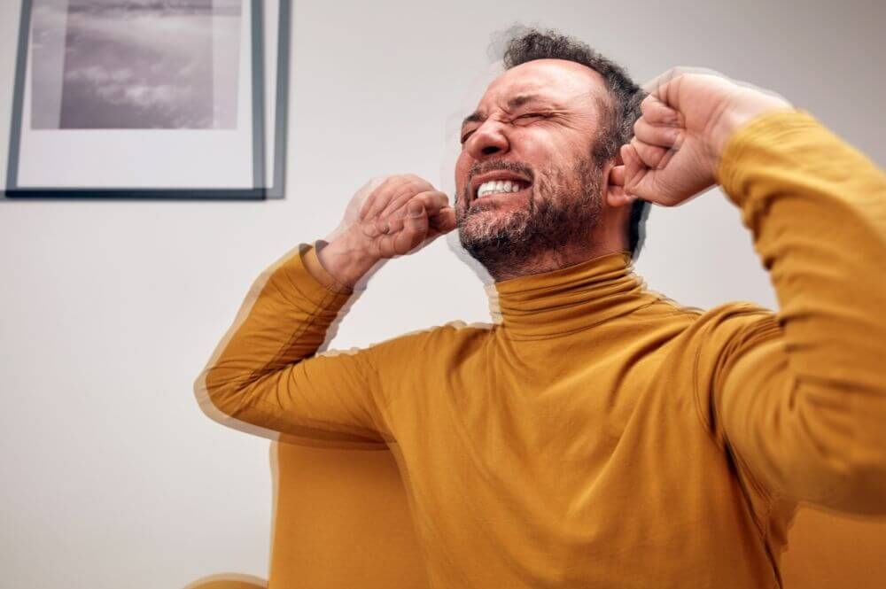 A man in a yellow turtleneck sits on a couch, grimacing and covering his ears with his hands, clearly distressed. A black-and-white photo hangs on the wall behind him.