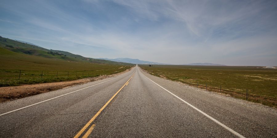 Open road stretching into horizon under blue sky, flanked by green fields, conveying freedom and adventure.