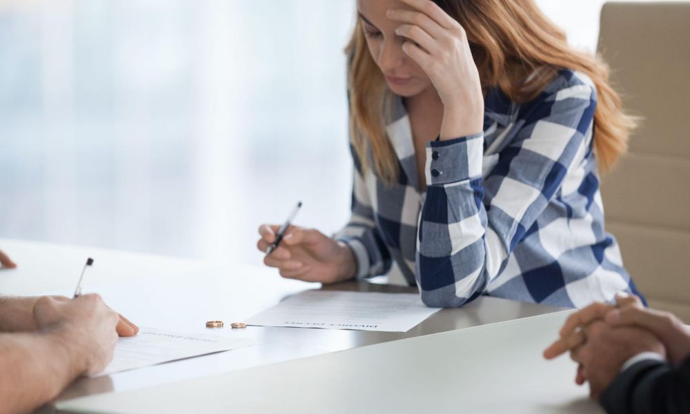 Woman signing divorce papers with rings on table, appearing stressed.