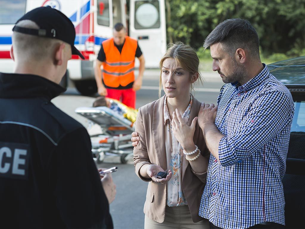 Couple talks to police officer after car incident, ambulance and paramedic in background assisting another person.