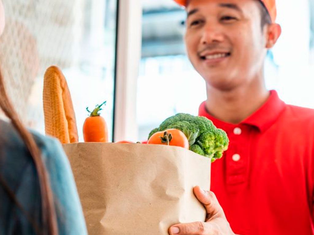 Delivery person handing over a grocery bag filled with fresh vegetables and bread to a customer.