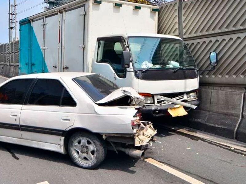 Car and truck collision on highway, damage visible on car rear and truck front, by roadside barrier.