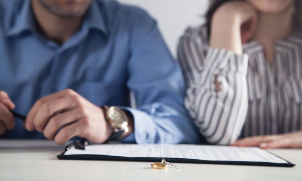 Two people, likely a couple, sitting at a table with wedding rings prominently displayed, suggesting a scenario involving marital commitment or dissolution. They appear somber and are not interacting.