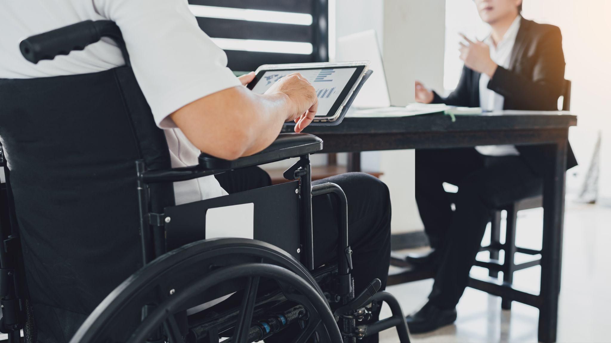 Person in a wheelchair using a tablet during a business meeting with a colleague at a desk.