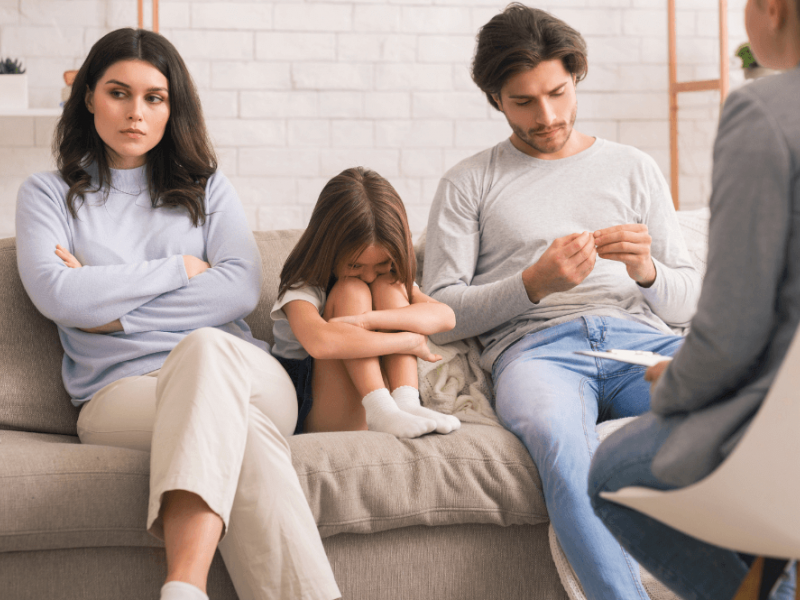 A family sits on a couch during a counseling session; a woman looks upset, a man appears contemplative, and a child rests her head on her knees; a therapist takes notes nearby.