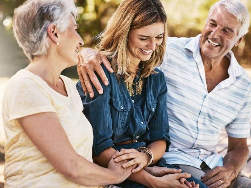 Two older adults and a younger woman sit close together, smiling and sharing a moment of affection outside on a sunny day.