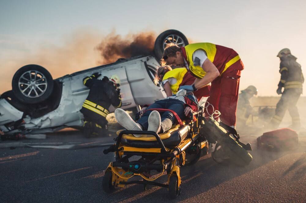 A paramedic attends to an injured person on a stretcher against the backdrop of an overturned, smoking vehicle with responders nearby.
