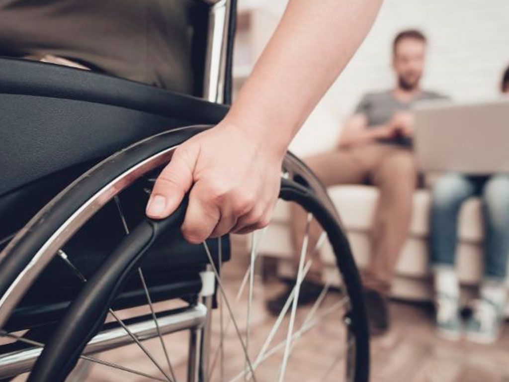 Close-up of a hand on a wheelchair wheel, with two people in the background sitting on a sofa.