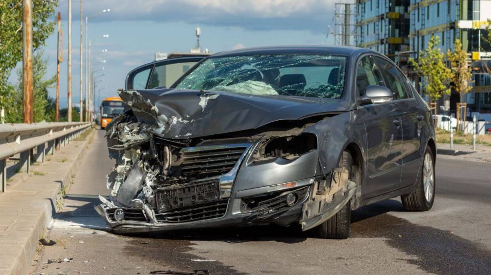 A damaged car with a crumpled hood and shattered windshield is stationary on a road near a construction site and a waiting bus.