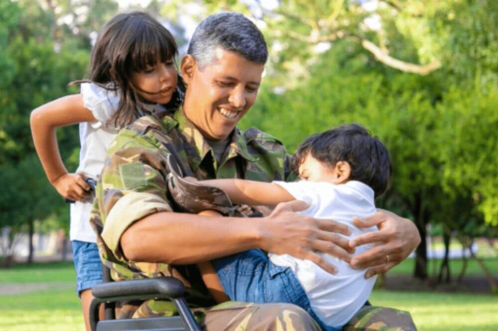 A smiling man in military uniform sits in a wheelchair, hugging two young children in a park. The children, a boy and a girl, are joyfully interacting with him, symbolizing family support and resilience. The lush green background suggests a peaceful outdoor setting in Norfolk, Virginia.