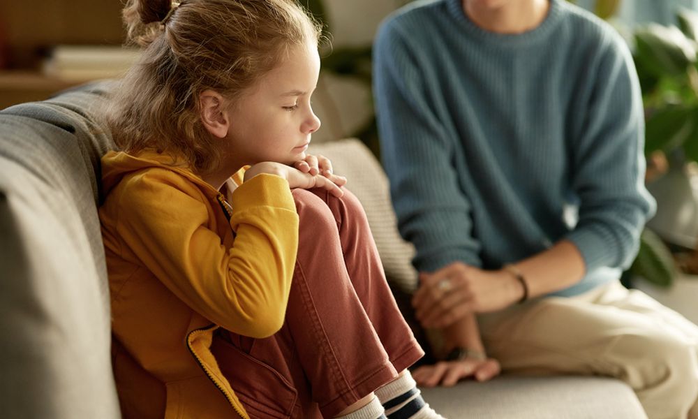 Child in a yellow jacket sitting on a couch beside an adult, looking thoughtful or concerned.
