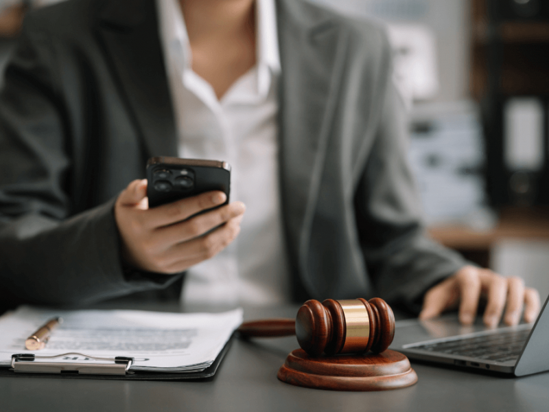 A person in a suit multitasks with a smartphone and laptop, near a gavel on a desk, possibly suggesting a legal setting.