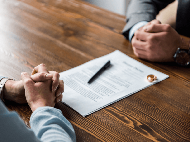 A document with text lies on a wooden table, while two hands clasped in contemplation face each other, suggesting a serious discussion or negotiation in a professional setting. A pen rests nearby.