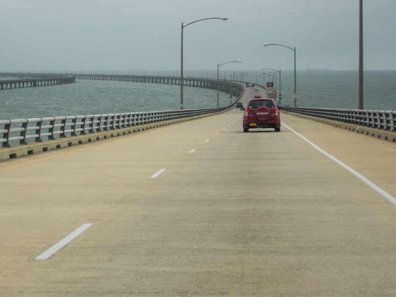 A view of the Hampton Roads Bridge-Tunnel with a red car driving along the bridge section on an overcast day. The bridge spans across a wide body of water, featuring safety barriers on either side and multiple lanes marked by clear white lines. In the distance, a parallel bridge section can be seen running alongside.