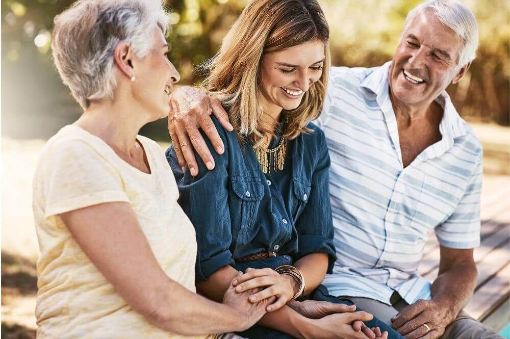 Two older adults and a younger woman sit close together, smiling and sharing a moment of affection outside on a sunny day.