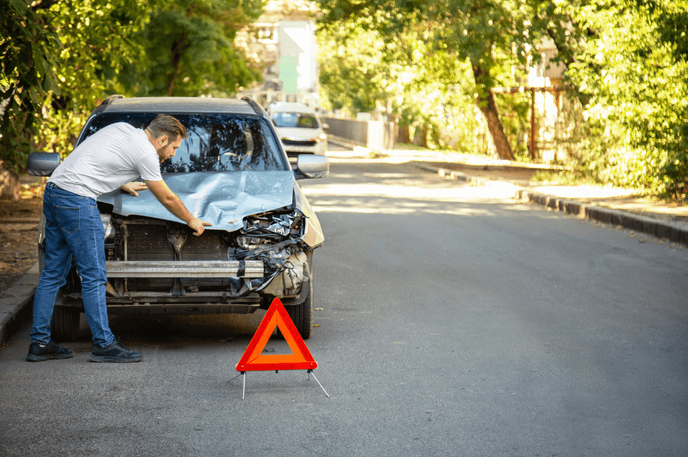 A man inspects a damaged car hood on a quiet, tree-lined street. A red warning triangle is placed on the road in front of the vehicle.