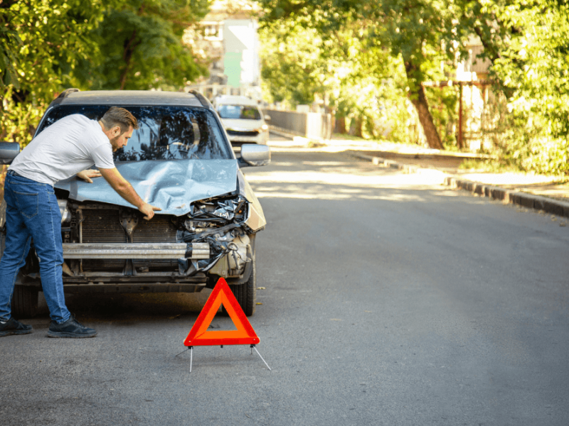 A man inspects a damaged car hood on a quiet, tree-lined street. A red warning triangle is placed on the road in front of the vehicle.