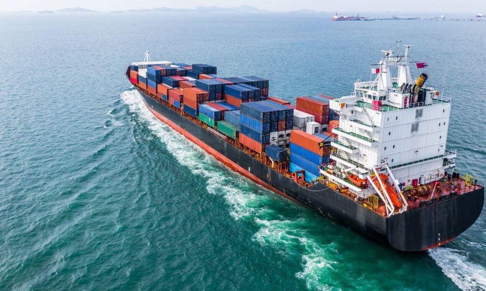 A large cargo ship carrying stacked shipping containers moves steadily across a vast blue ocean, with distant land visible on the horizon.