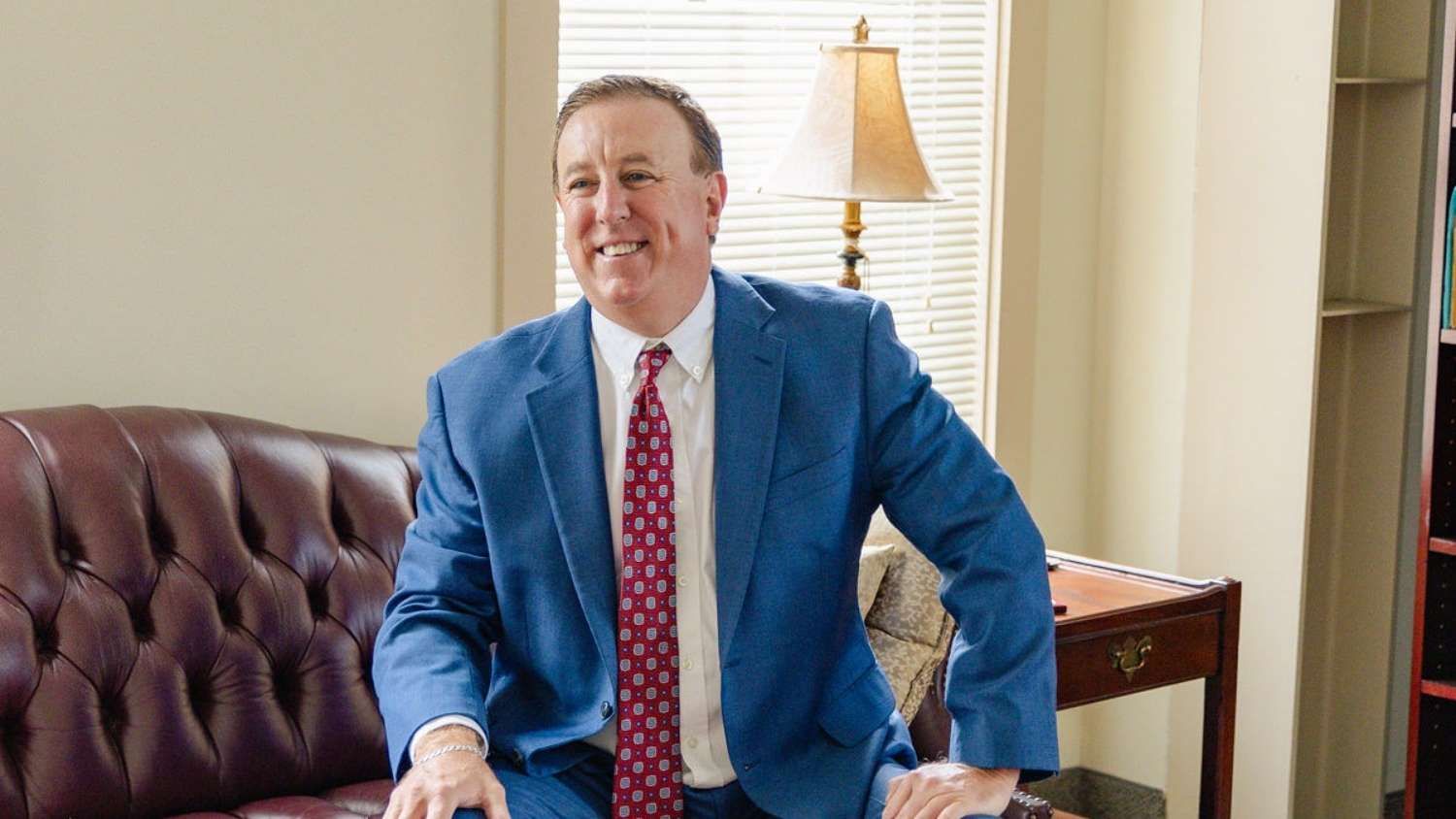 Smiling man in a blue suit sitting on a couch in an office with a lamp in the background.