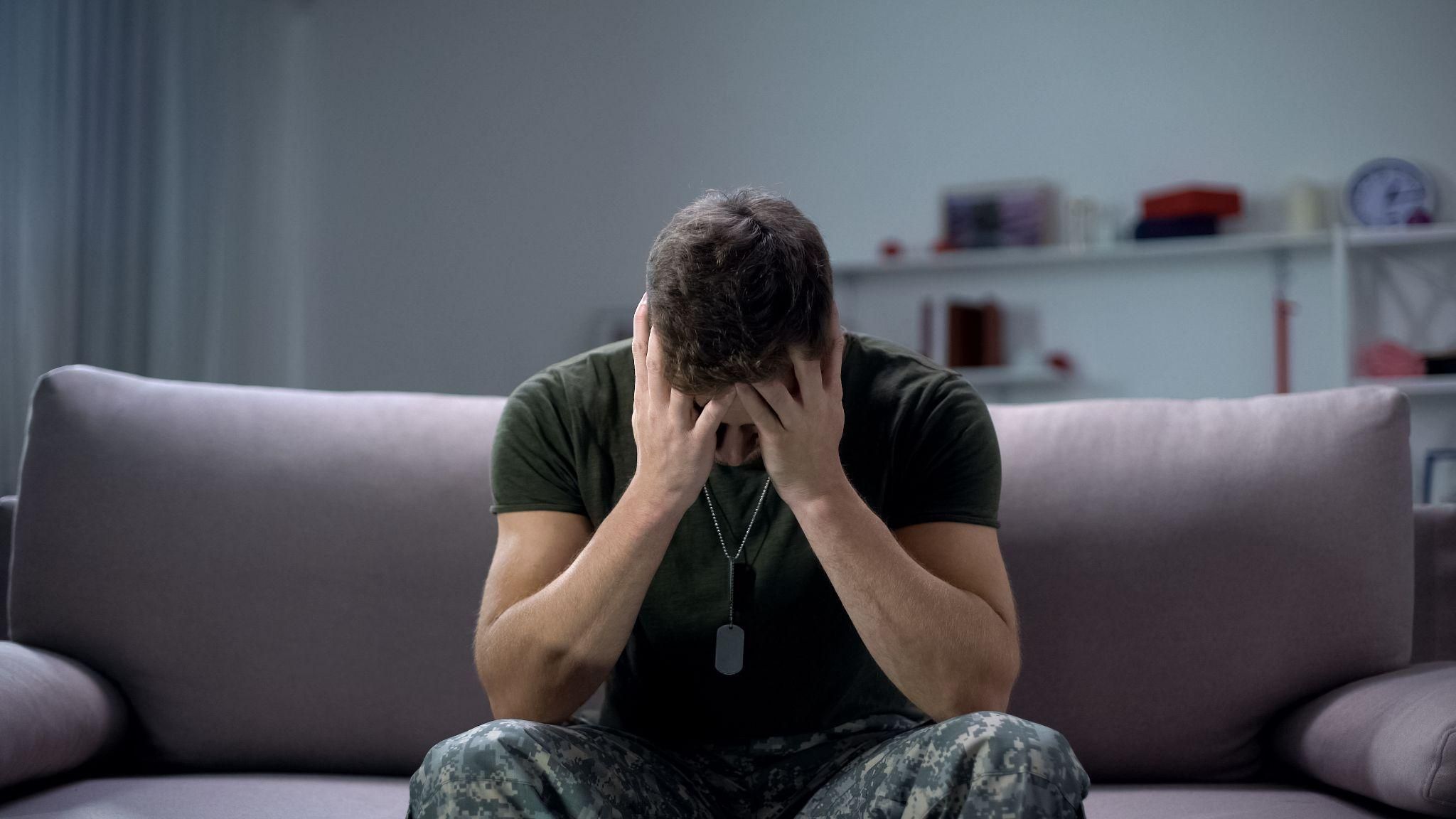 Man in military uniform sitting on sofa, hands covering face, representing stress or anxiety in a domestic setting.