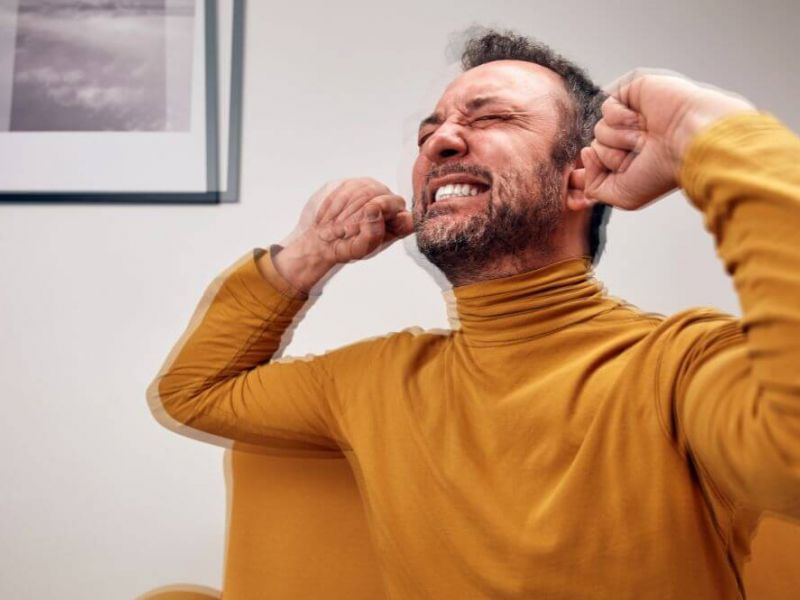 A man in a yellow turtleneck sits on a couch, grimacing and covering his ears with his hands, clearly distressed. A black-and-white photo hangs on the wall behind him.