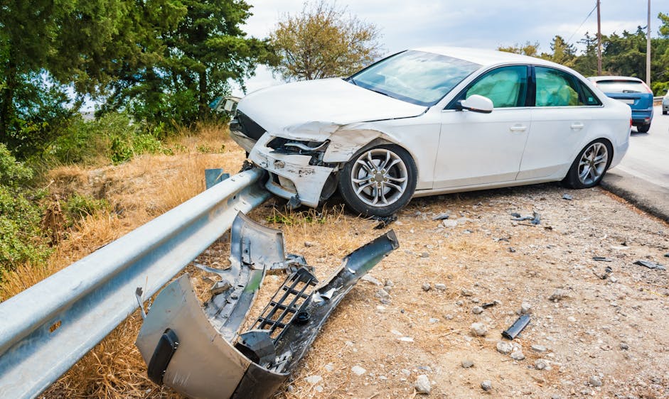 A damaged white car is perched on a guardrail beside a road, with wreckage scattered on the ground, indicating a recent collision in a rural setting.