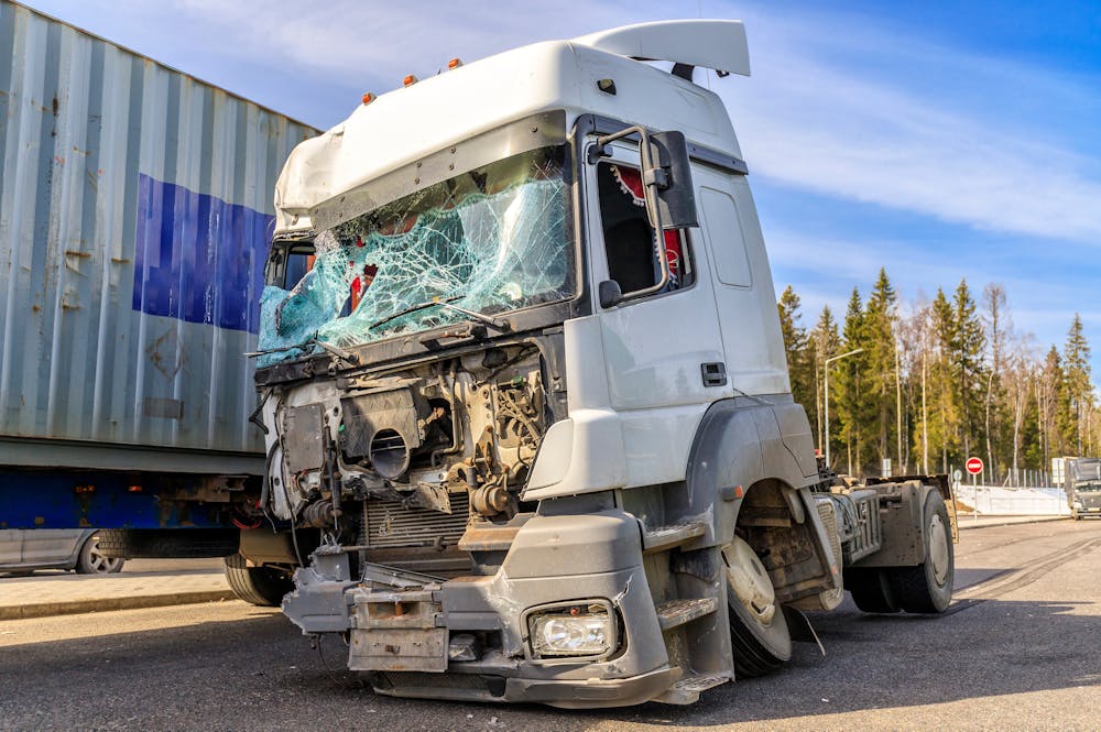 Damaged truck with shattered windshield after accident near shipping container on sunny day.