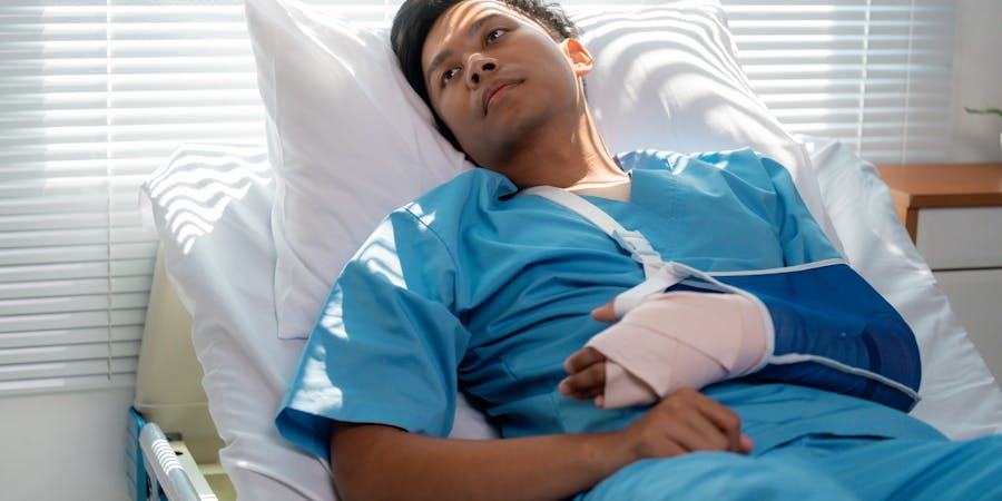 Person in a hospital bed with arm in a blue sling, looking contemplative in a brightly lit room.