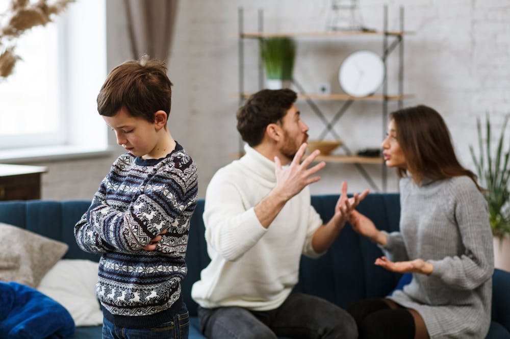 Boy looking sad while a couple argues in the background on a sofa in a modern living room.