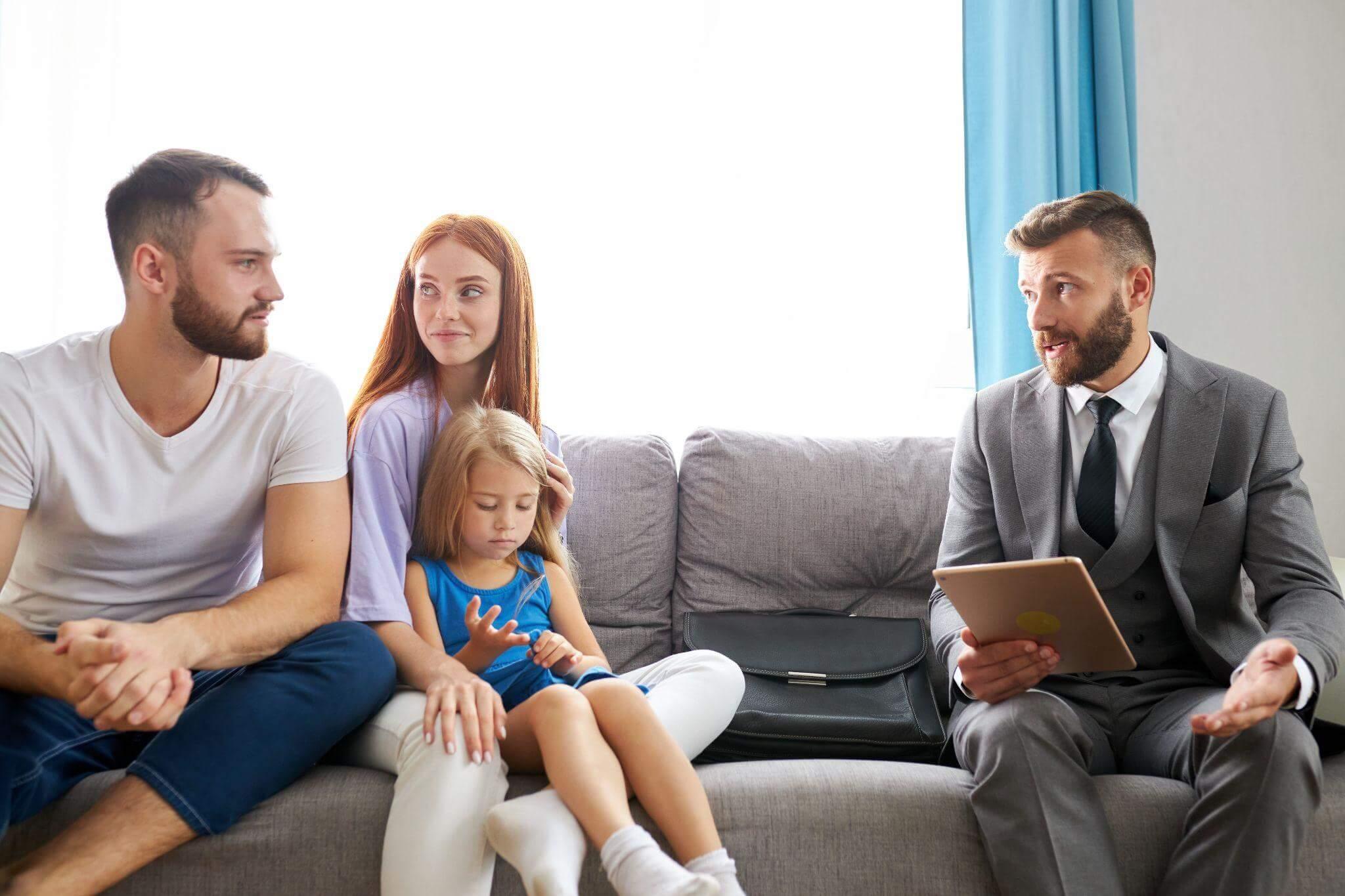 A family of three, a man, woman, and child, sits on a sofa while a man in a suit, holding a tablet, talks to them in a bright room.