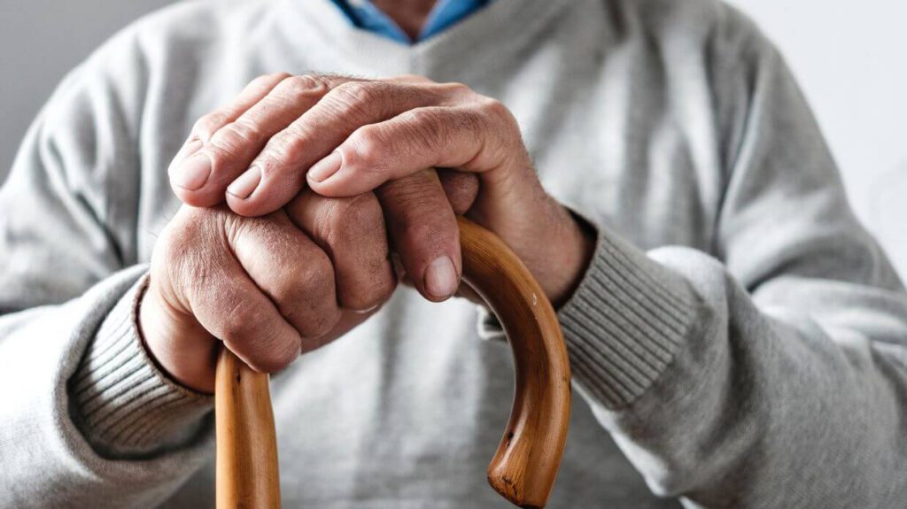 Close-up of an elderly person's hands resting on a wooden cane, symbolizing the support and stability provided by Supplemental Security Income (SSI) services. The person is wearing a grey sweater, indicating a sense of comfort and security in their daily life.