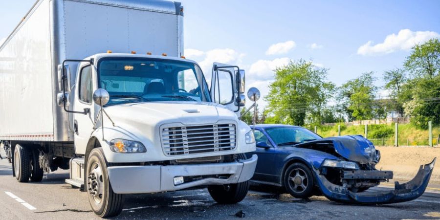 A truck accident scene in Newport News, Virginia, showing a white truck and a black car with significant front-end damage. Both vehicles are stationary on the road, with clear skies and trees in the background.