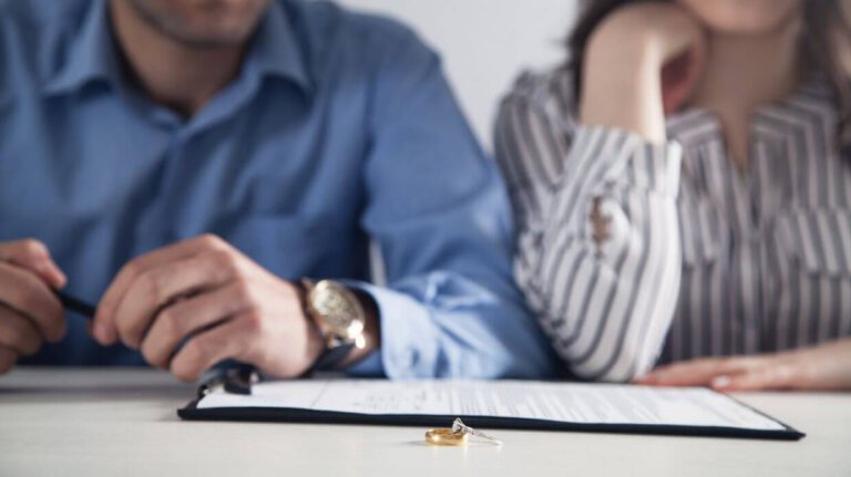 Two people, likely a couple, sitting at a table with wedding rings prominently displayed, suggesting a scenario involving marital commitment or dissolution. They appear somber and are not interacting.