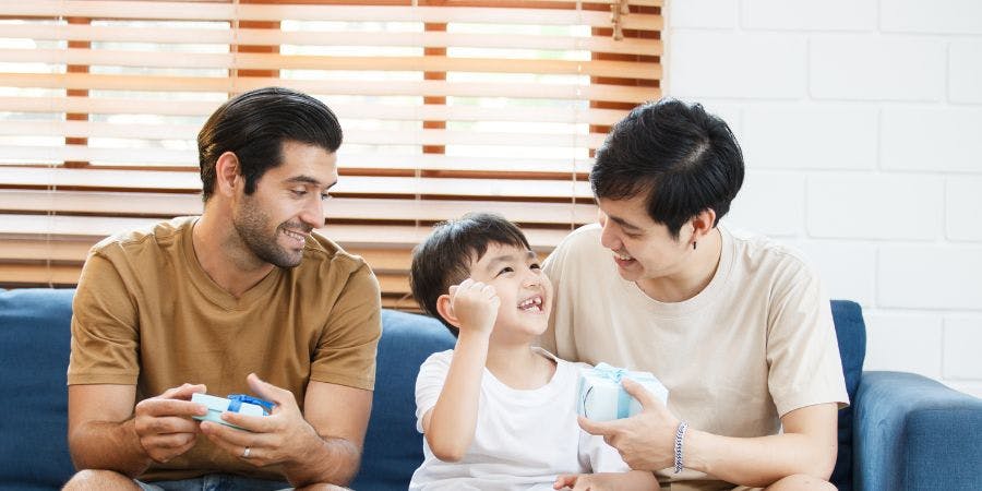 Two adults and a child are sitting on a couch, smiling and playing with video game controllers; behind them is a window with blinds.