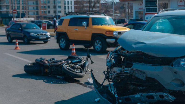 Traffic accident scene with a damaged car and a motorcycle, surrounded by traffic cones, in an urban area.