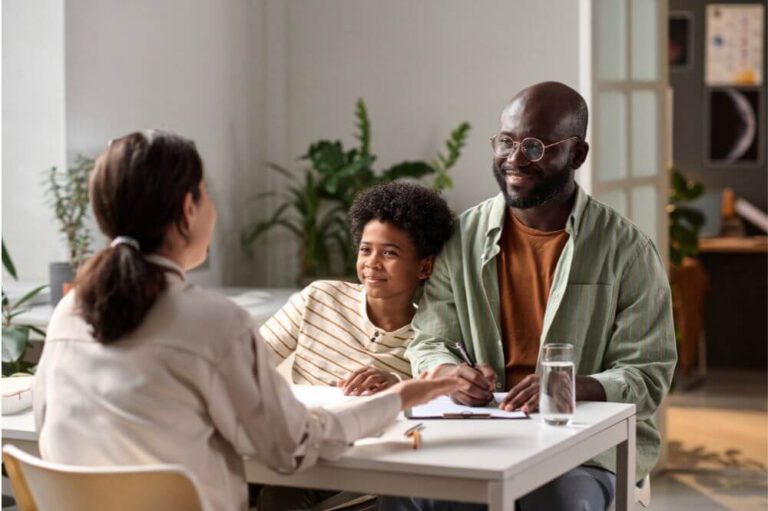 A man and a young child smiling, sitting at a table during a meeting with a woman in an indoor, plant-adorned setting.