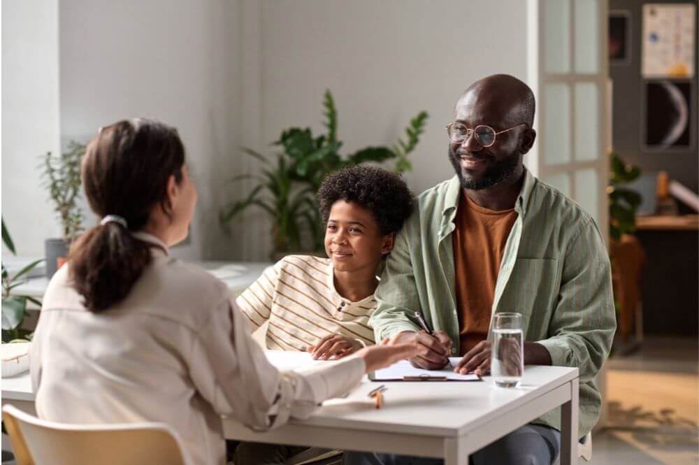 A man and a young child smiling, sitting at a table during a meeting with a woman in an indoor, plant-adorned setting.